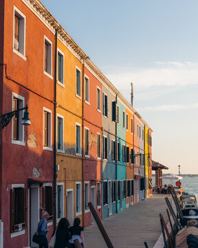 View of vibrant, sun-kissed buildings casting long shadows along the waterfront promenade, a picturesque scene of everyday life unfolding, Burano, Veneto, Italy.