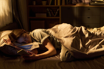 Teenage girl lying in bed using digital tablet at night, resting head on pillow while holding device in one hand, illuminated by screen light, bookshelf in background © Mediaphotos