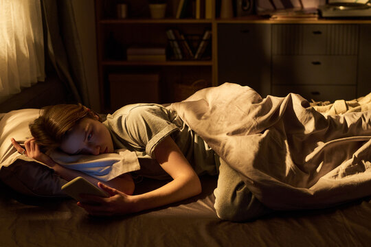 Teenage girl lying in bed using digital tablet at night, resting head on pillow while holding device in one hand, illuminated by screen light, bookshelf in background