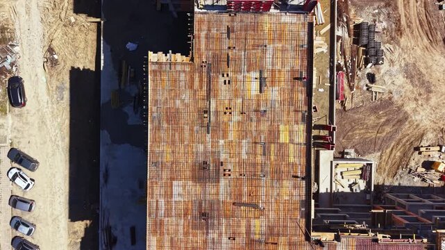 Workers are installing steel beams on a construction site. Vehicles are parked nearby. Building materials are scattered around the area. The site is active with ongoing work tasks.