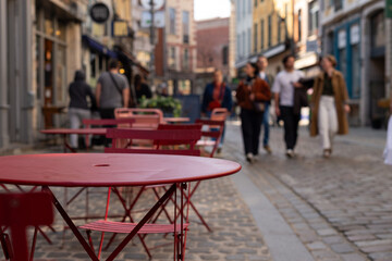 Red metal tables and chairs at an outdoor cafe on a cobblestone street in Lille, France, with blurred people walking in the background. © dsbedarieva