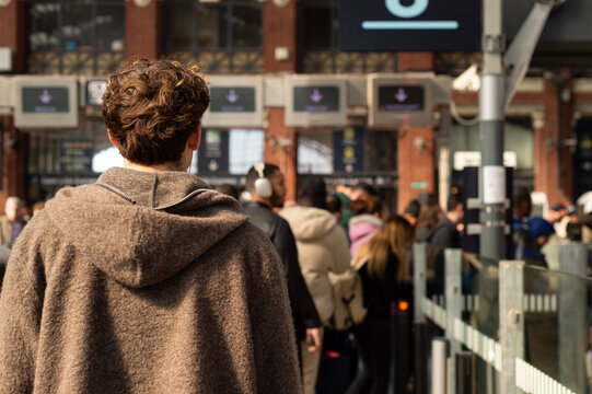 Rear view of a man with curly hair wearing a brown hoodie standing in a crowded train station in Lille, France. Electronic departure boards are visible in the background.