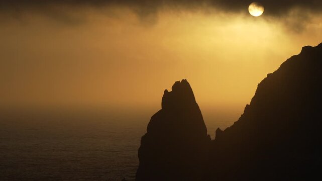 Sunset coastal cliffs dramatic sea stack silhouettes against a golden sky and misty ocean as the sun peeks through dark clouds