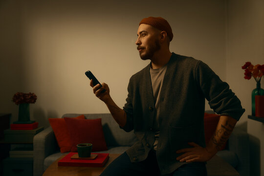 A stylish, tattooed man in an orange beanie and cardigan sits in a warm, dimly lit living room, holding a smartphone while looking pensively away. Cozy, cinematic interior with a moody atmosphere.