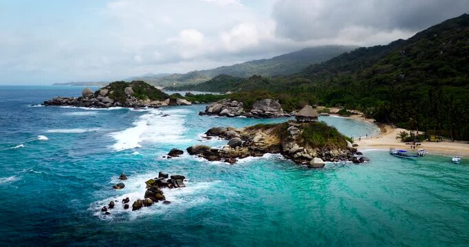 Aerial footage over Tayrona National Park, Colombia, revealing a turquoise cove, rocky islets, white surf and tropical shoreline backed by lush forested hills