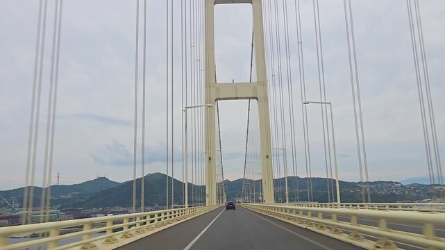 Driving on Hakucho Bridge in Muroran, Hokkaido, Japan, seeing the main towers and suspension cables with mountainous terrain in the background on an overcast day