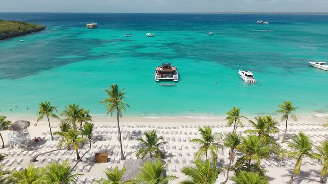 Aerial view of tropical beach in Dominican Republic with turquoise water, anchored boats, palm trees and white sand resort shoreline under clear sunny skies. Wide shot.