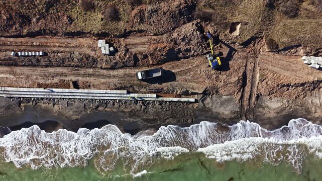 Workers are seen operating machinery and laying materials along the shoreline. Waves crash on the beach as construction progresses on a building project.