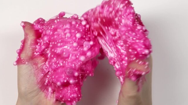 Playing with textured pink slime toy with bubbles, stretching the gooey substance for fun and stress relief. Close up and top view of female hand holding pink shining slime, squeezing it.