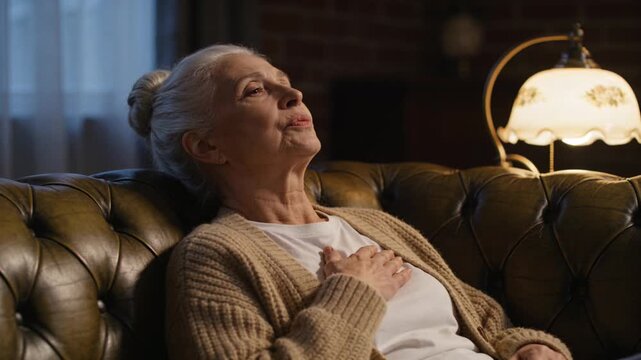Taking deep breaths after sitting, Senior woman holding chest on tufted sofa, wearing cardigan