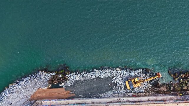 Workers are using a machine to move rocks and sand along the shore next to blue water. The scene shows a construction area and building materials present on the shoreline.