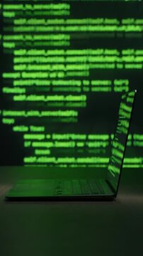 Laptop with hacking software on table in dark room, green code lines moving in background. Cyber attack, malware activity, data breach and cybersecurity threat concept. Vertical video. Copy space