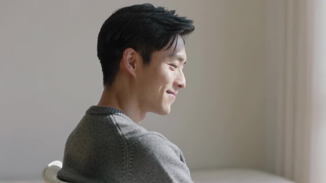 Glancing toward window, Asian man wearing grey sweater softening and smiling in softly lit room