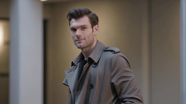 Turning toward camera after noticing it, man in grey trench coat offering closed-mouth smile, lobby