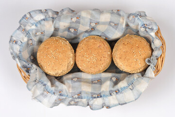 Three golden brown whole grain bread rolls topped with quinoa seeds arranged in a wicker basket with a blue checkered cloth