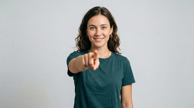A smiling woman in a dark teal t-shirt points her finger directly at the viewer a soft grey studio background, conveying a direct and friendly message.