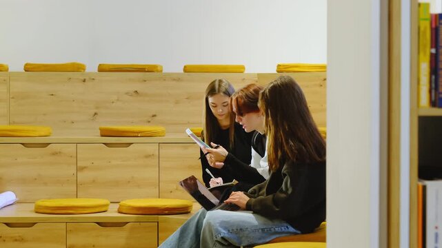 Teen study trio sits on wooden amphitheater steps reading worksheet pages. Teen boy holds pen explaining point while teen girls write notes nearby