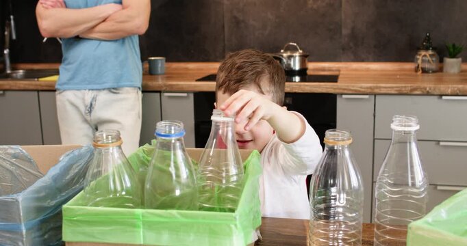 Young boy lifts empty plastic bottle above green recycling box in kitchen. Child sorts clear container with hand while father watches waste separation task nearby
