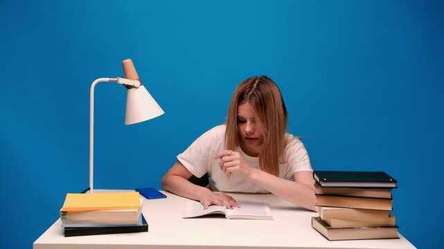 Tired female student waking up and studying hard at her desk with a stack of books and a lamp against a blue background