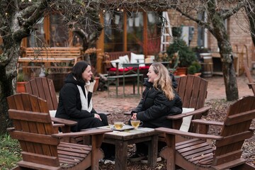 Obraz premium Two pretty caucasian women in their 40s wearing warm clothes spending time outdoors in autumn park cafe. Mature females enjoying personal time, talking, drinking tea, relaxing and breathing fresh air.