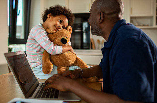 Father working on laptop while child hugs teddy bear at kitchen table
