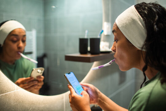 Woman brushing teeth while using smartphone in bathroom mirror
