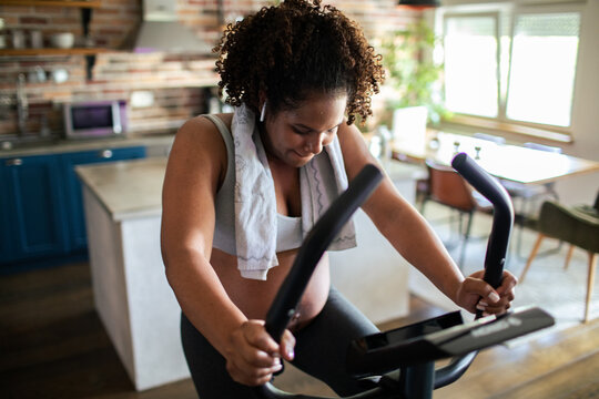 Pregnant woman cycling on exercise bike at home kitchen