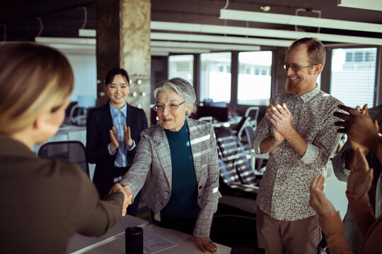 Coworkers applaud as senior woman shakes hands in modern office