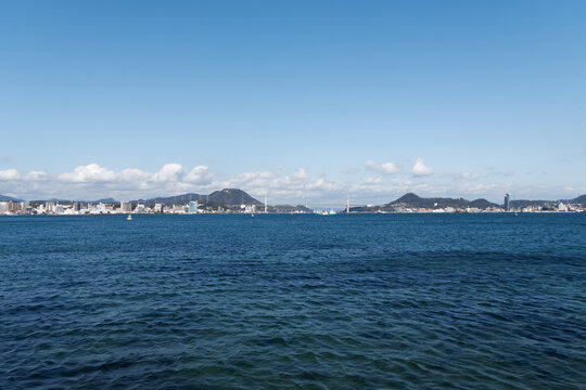 Distant panoramic view of the Kanmon Bridge over the blue sea of Kanmon Strait under a clear blue sky with white clouds.