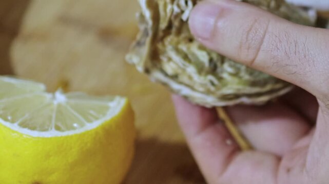 Man hand opens oysters at home on wooden surface using small knife. Lemon in frame, seafood preparation, kitchen scene, fresh ingredients, close view of shells and texture.