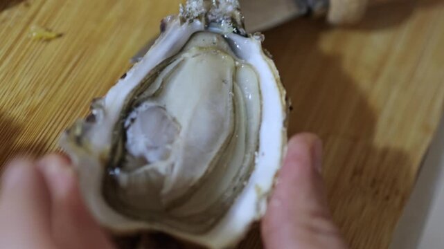 Man hand opens oysters at home on wooden surface using small knife. Lemon in frame, seafood preparation, kitchen scene, fresh ingredients, close view of shells and texture.