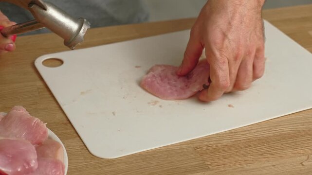 Person using meat tenderizer on raw chicken. Chef preparing pork loin slice with meat mallet on cutting board. Man tenderizing fillet. Cooking cutlets pounding poultry breast close-up. Culinary dish.