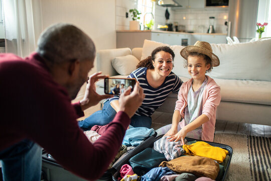 Father photographing mother and daughter packing suitcase at home