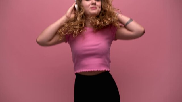 Happy young woman with curly hair dancing and listening to music with headphones on a pink background