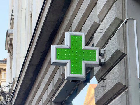 Green illuminated pharmacy cross sign mounted on a stone building facade in the city center. Healthcare wayfinding, medical retail identity, and urban signage in a historic streetscape context