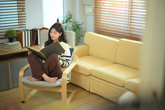 Happy relaxed young woman resting on armchair reading book and holding coffee cup