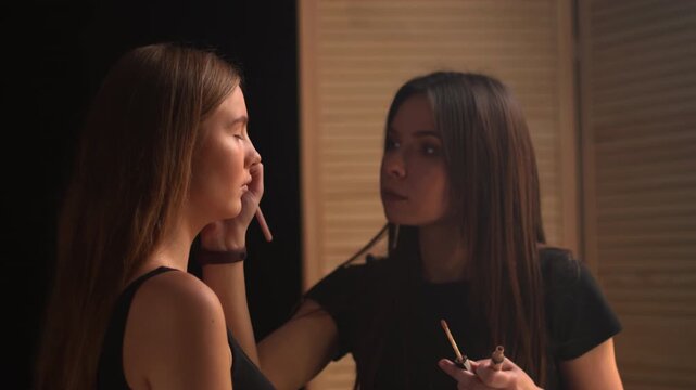 Professional makeup artist applying concealer and foundation with a brush to a young model's face in a beauty salon, highlighting the cosmetic procedure and skincare concept