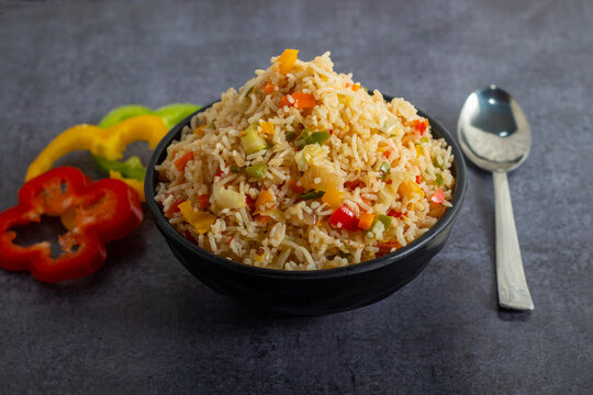 Black bowl filled with vegetable fried rice, colorful peppers around, served with spoon on dark textured background.
