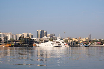 Naklejka premium 12.12.2025 Dubai, UAE. Traditional dhow and modern yacht at Dubai Creek in Deira with old district buildings and Dubai skyline in background. Contrast of heritage and modern city.