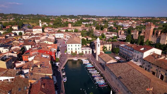 Italy Lake Garda aerial drone fly-out from scenic canal with tower and boats to wide lake panorama. Reveal of Lazise promenade and Scaliger castle with gardens 4k travel destination scenery