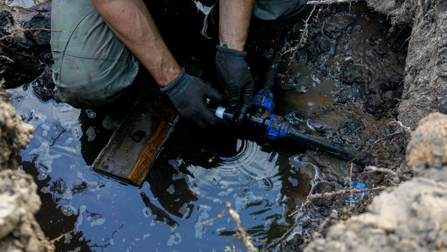 Gloved plumber crouching in a flooded muddy trench replacing a blue compression pipe, fitting by hand during an underground water leak repair on residential property.