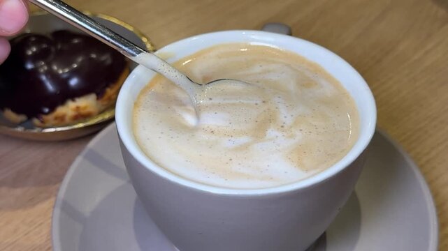 spoon stirring cappuccino on wooden table with chocolate eclair on gray plate. Cozy cafe atmosphere, coffee ritual, dessert pairing and warm lifestyle moment.
