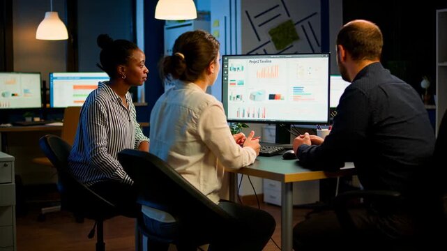 Speaking left woman wearing striped top guiding team on charts at office desk reviewing timeline
