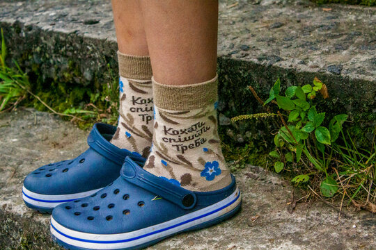 teenage legs in blue crocs and floral socks with inscription "love must hurry", brown-cream, nature, hiking, off-season, hike, childrens camp, excursion, closeup, view straight, forest area