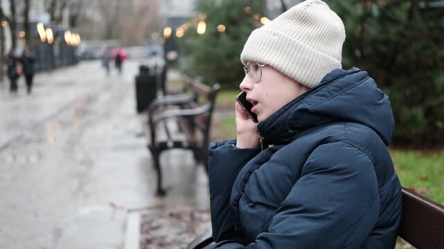 Teenager Girl Walking Alone on City Street with Smartphone. Portrait of 14 years old girl wearing eyeglasses, wool hat, winter jacket using mobile phone. Urban Style. Rainy mood weather. Cold season