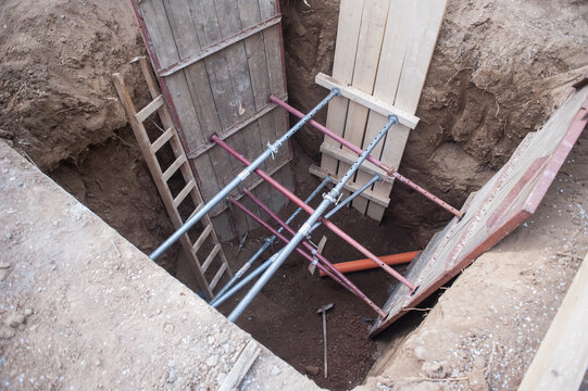 Workers prepare for a major construction project, digging deep into the earth while ensuring safety with sturdy shoring and ladders. Surrounding soil creates a rugged scene of labor