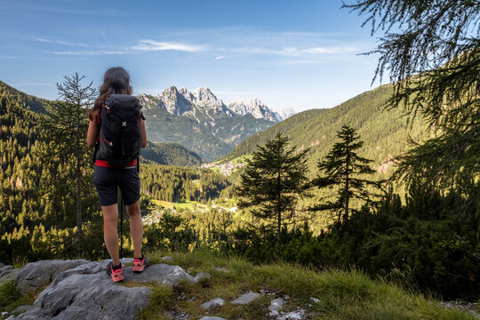 Travel, tourism: Woman hiker with backpack looks at mountain landscape, forest and village. Volaia lake, Italian-Austrian border, Friuli Venezia Giulia.