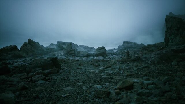 Mistshrouded rocky coastline at dusk, low light and cold atmosphere with scattered pebbles and jagged boulders, windblown surf and distant muted horizon evoke