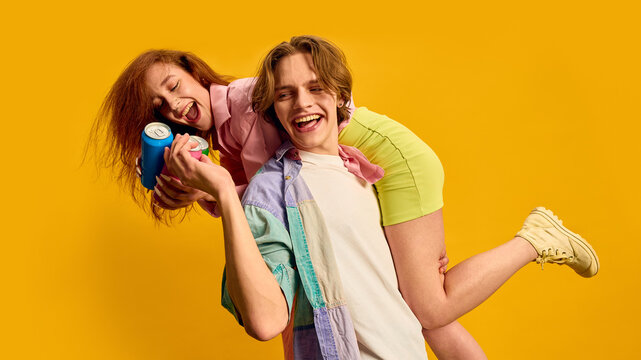 Young man carrying smiling woman on shoulder holding drink cans on bright yellow background. Concept of Gen Z lifestyle, relationship bonding, beverage promotion, fun youth marketing.