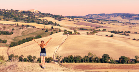 Naklejka premium Young woman with open arms enjoying the golden sunset over the rolling hills of Tuscany, Italy. Breathtaking landscape captures the iconic rolling hills of Val d'Orcia during the golden sunset